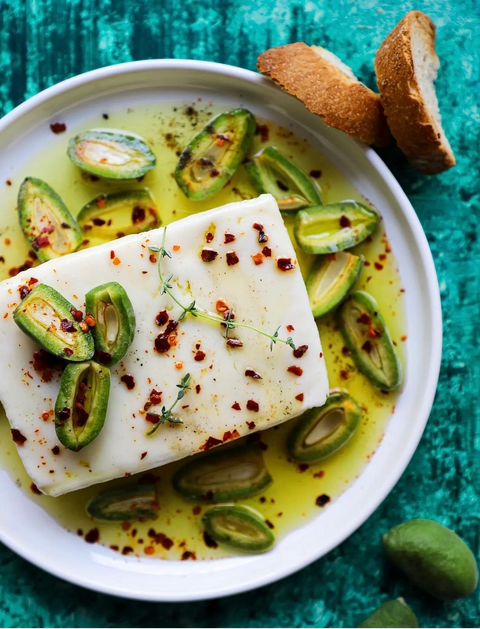 Feta-Käse mit Minos Nativem Olivenöl Extra beträufelt, garniert mit geschnittenen grünen Oliven, Chiliflocken und frischem Thymian, serviert mit knusprigem Brot.