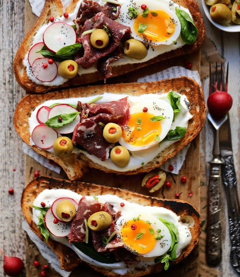 Frisch zubereitete Stullen mit Minos BIO Grüne Oliven mit Paprika, Rindfleisch, Spinat, Radieschen und einem perfekt gekochten Ei, serviert auf rustikalem Holzbrett, ideal für einen mediterranen Genuss.
