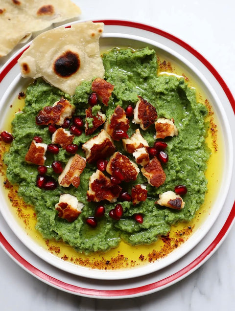 Ein appetitlicher Teller mit grünem Olivenpüree, garniert mit gegrilltem Käse und Granatapfelkernen, serviert mit frisch gebackenem Brot. Die Minos Bio Oliven ohne Stein sind eine perfekte Ergänzung zu diesem gesunden und leckeren Gericht, das mit hochwertigem Olivenöl verfeinert ist.