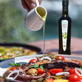 Person gießt Minos natives Olivenöl Extra aus einer Flasche über eine mediterrane Gemüsepfanne, die mit frischen Zutaten und Oliven garniert ist.